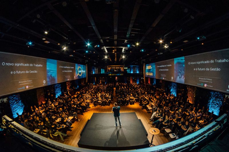 Presenter on stage in a conference venue with audience and projection screens
